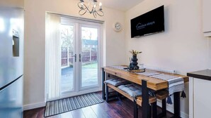 Una cucina moderna con un tavolo in legno, sedute a banco, un lampadario, una televisione a schermo piatto e una porta scorrevole in vetro che dà su un giardino.