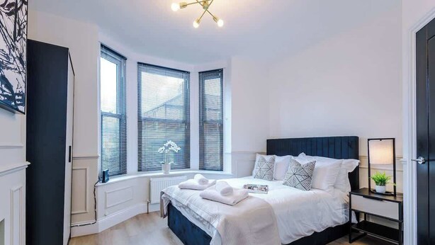 Quarto com cama, mesa de cabeceira, luminária e janelas.