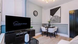Uma sala de estar moderna com uma TV de tela plana, uma mesa de jantar com cadeiras, um relógio na parede e uma obra de arte emoldurada.
