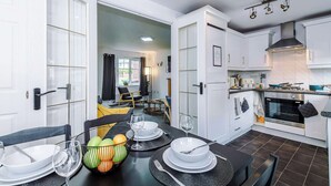A modern kitchen with white cabinets, a stove, and an oven. There is a dining area with a table set for two, including plates, glasses, and a fruit bowl. - Sleeps 5 Near Manchester Centre Free Parking (Greater Manchester)