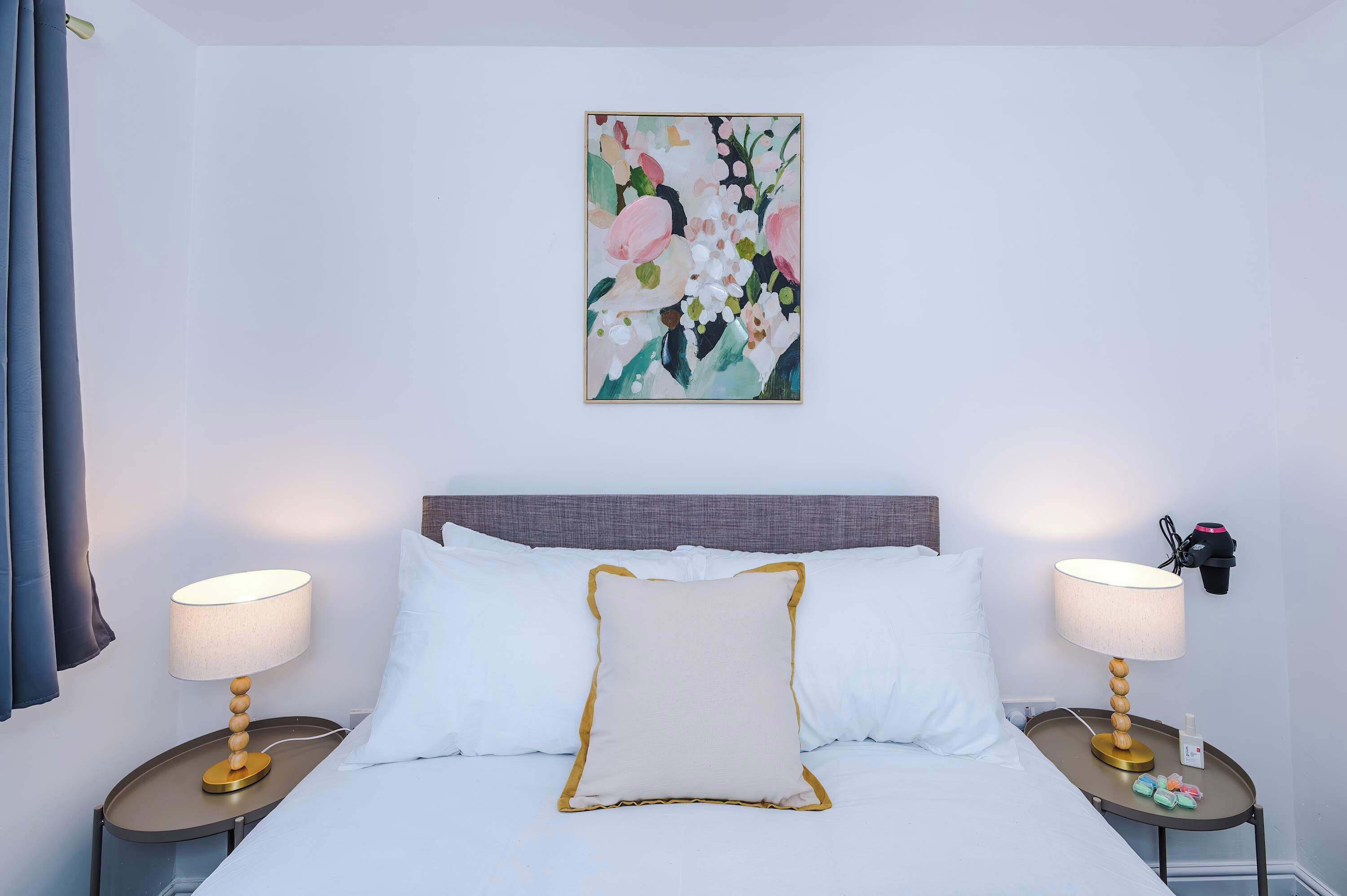 A hotel room with a bed, two bedside tables with lamps, a wall-mounted artwork, and a nightstand with a lamp and a phone.