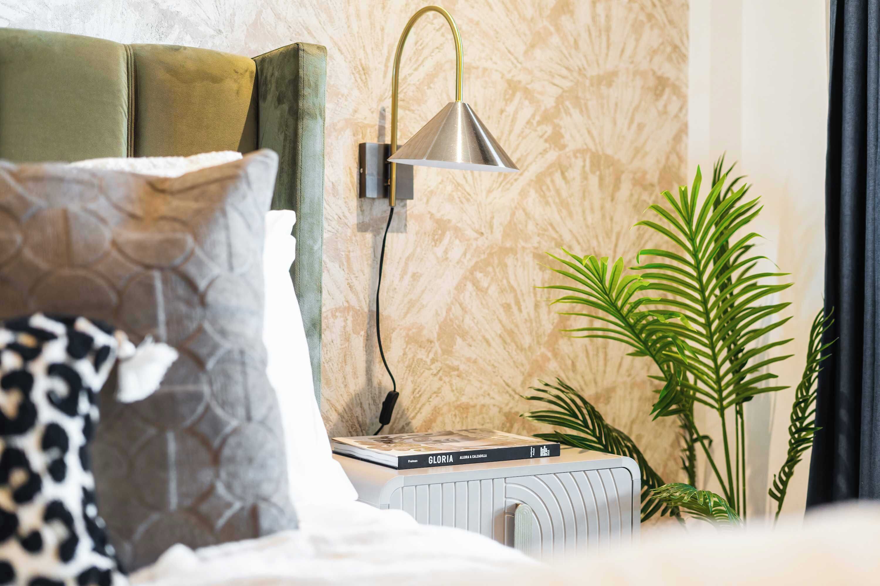 Ein ordentlich eingerichtetes Schlafzimmer mit einem Bett, Kissen, einem Nachttisch mit Büchern, einer Wandlampe und einer Zimmerpflanze.