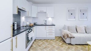 Eine moderne Küche mit weißen Schränken, einer schwarzen Arbeitsplatte und goldenen Griffen. Im angrenzenden Wohnbereich befindet sich eine Couch mit gemusterten Kissen.