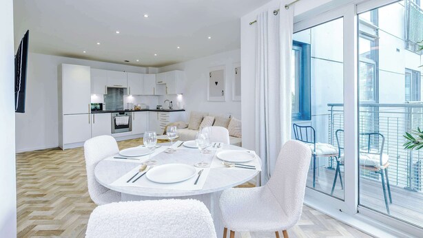 Eine moderne Küche mit weißen Schränken, ein Essbereich mit rundem Tisch für vier Personen und ein Balkon mit blauen Stühlen.