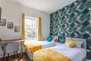 Une chambre avec deux lits, un bureau et une chaise. Il y a une fenêtre avec des rideaux jaunes, et les murs sont décorés d’un motif géométrique.