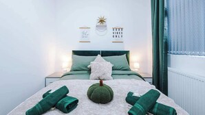 Ein ordentlich gemachtes Bett mit grüner Bettwäsche, ein kleiner Dekopumkin und zusammengerollte Handtücher auf einer weißen Oberfläche.