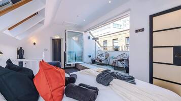 A modern hotel room with a bed, pillows, a small table, a chair, and a balcony with a view.