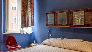 Quarto com cama, uma cadeira vermelha, janela com cortinas estampadas e prateleira fixa com cestos trançados.