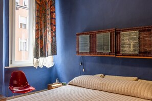 A room with a bed, a red chair, a window with patterned curtains, and a wall-mounted shelf with woven baskets. - GuestHost - Stylish Apartment in the Heart of Livorno (Livorno)