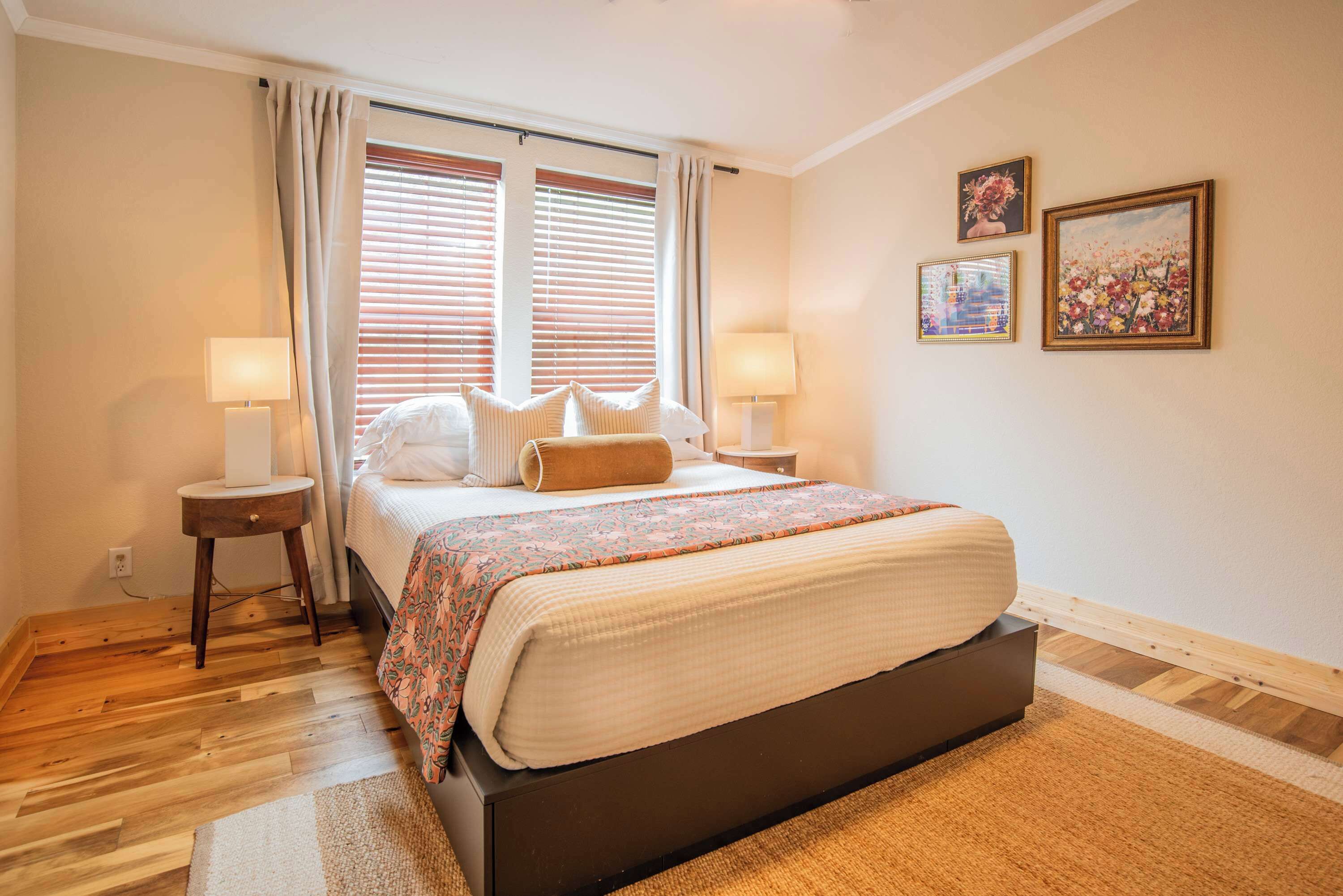 Une chambre à coucher comprenant un lit, deux tables de chevet, une fenêtre avec des stores et trois tableaux encadrés accrochés au mur.