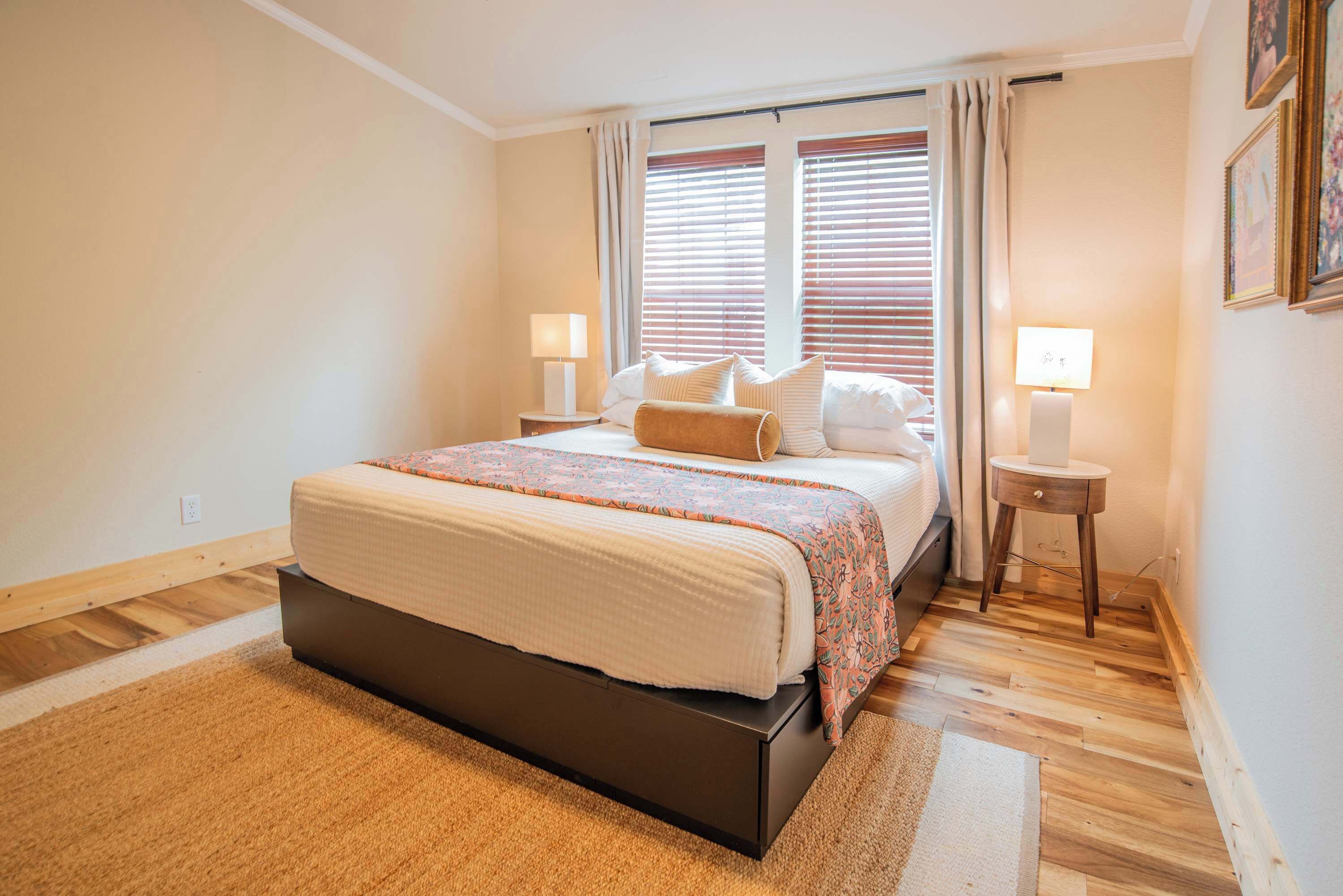 Un dormitorio con una cama, dos mesitas de noche, una ventana con persianas y una lámpara de pared.