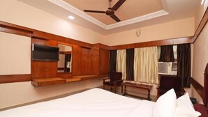 A hotel room with a large bed, a ceiling fan, a flat-screen TV, a wooden bench, and a window with curtains.