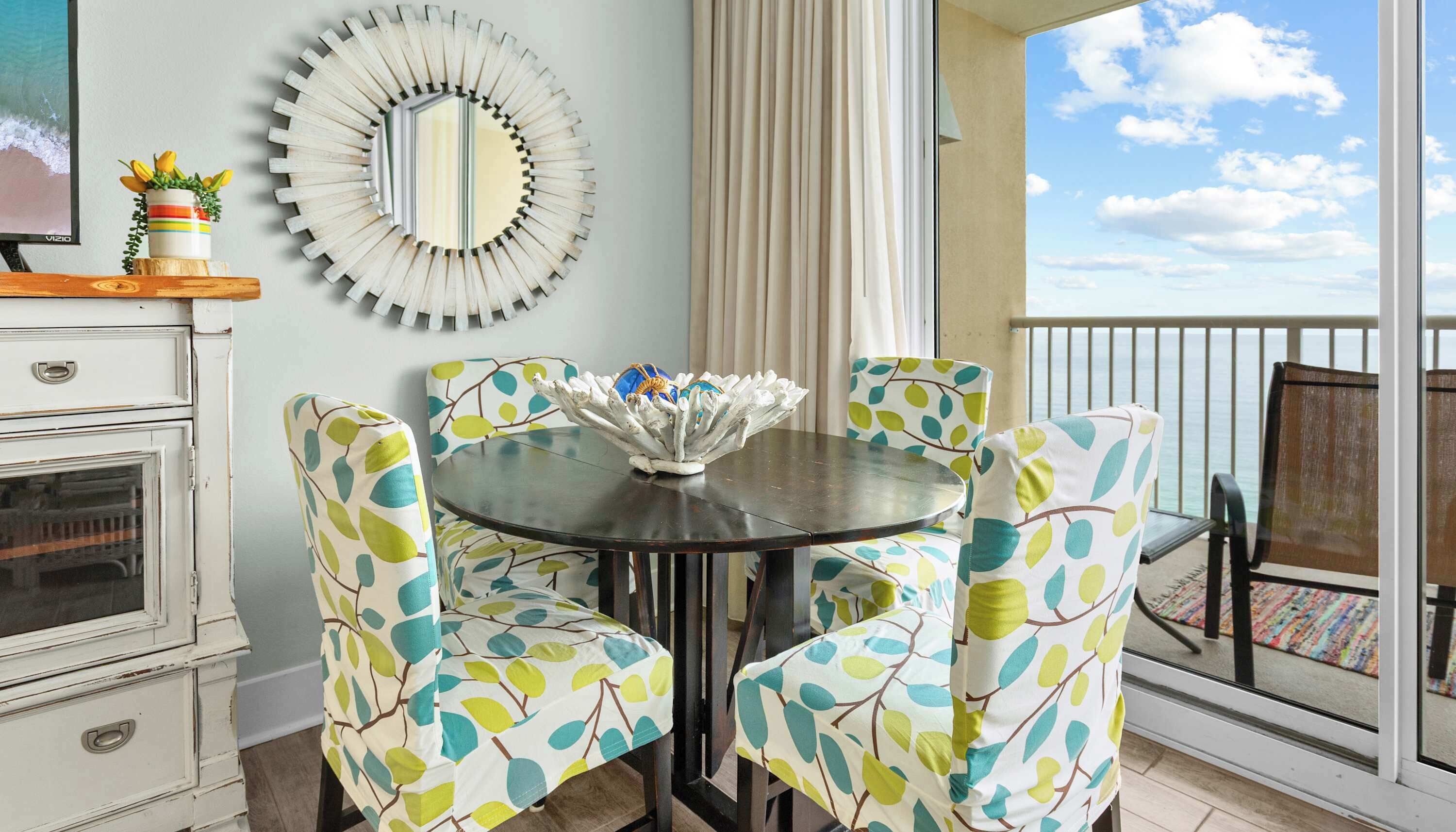 Un comedor con una mesa redonda, sillas con motivos de hojas, un espejo decorativo y vista al océano.