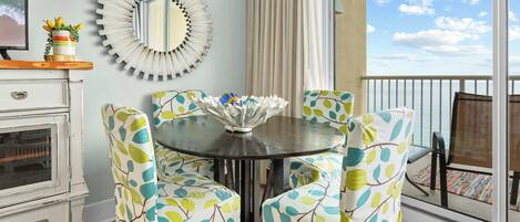 Un comedor con una mesa redonda, sillas con motivos de hojas, un espejo decorativo y vista al océano.