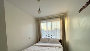 Een eenpersoonsbed met wit beddengoed in een kamer met een raam, een plafondlamp en een haak aan de muur.