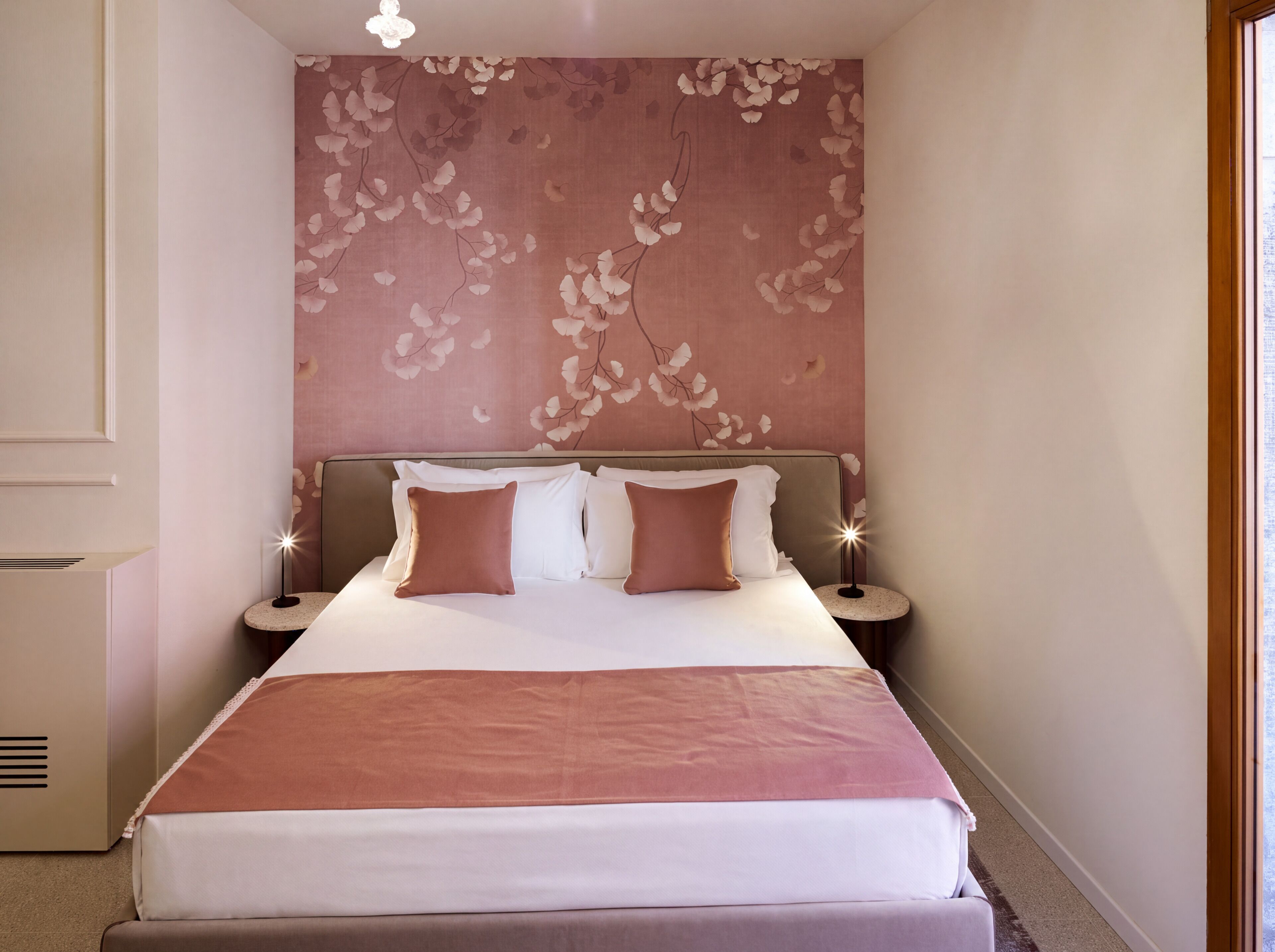 A neatly made bed with a pink and white color scheme, a wall with a floral pattern, and a small bedside table with a lamp.