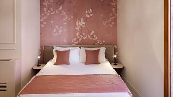 A neatly made bed with a pink and white color scheme, a wall with a floral pattern, and a small bedside table with a lamp.