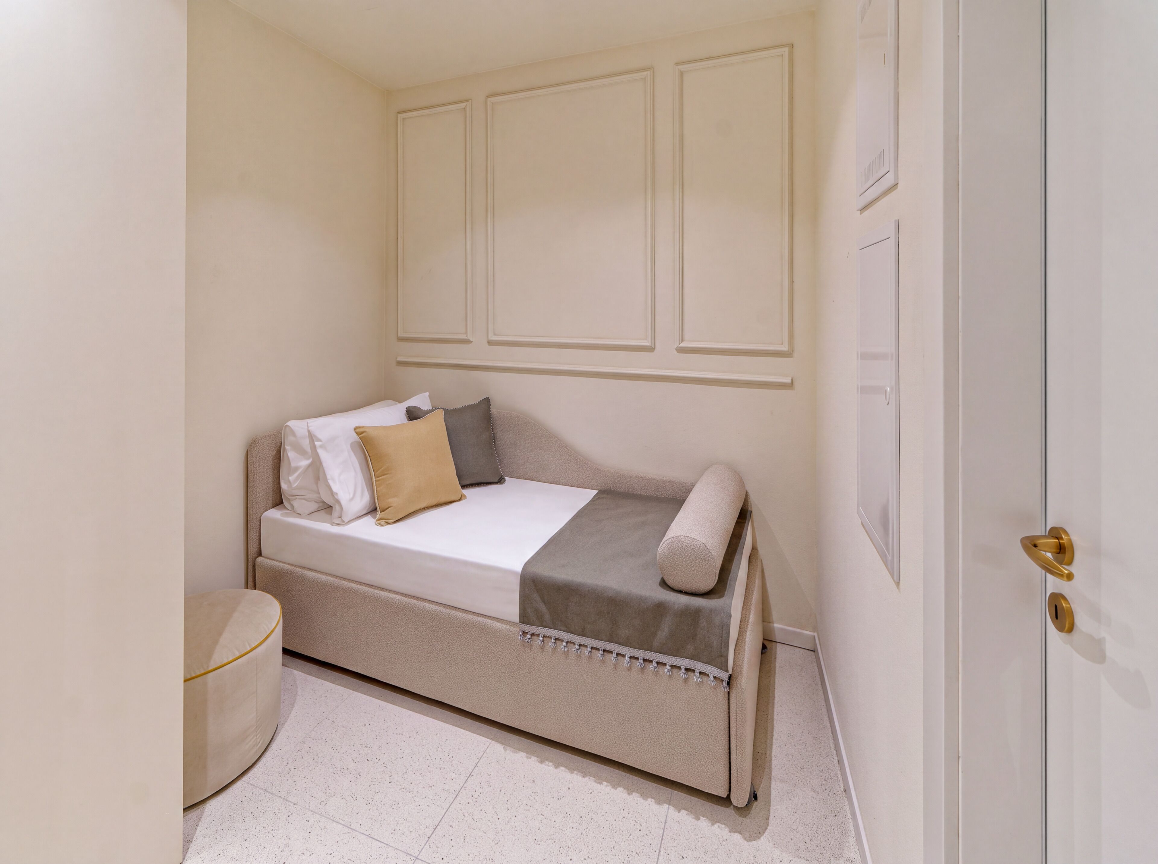 Una habitación pequeña con cama individual, pared blanca sencilla, puerta con pomo dorado y una mesita de noche.