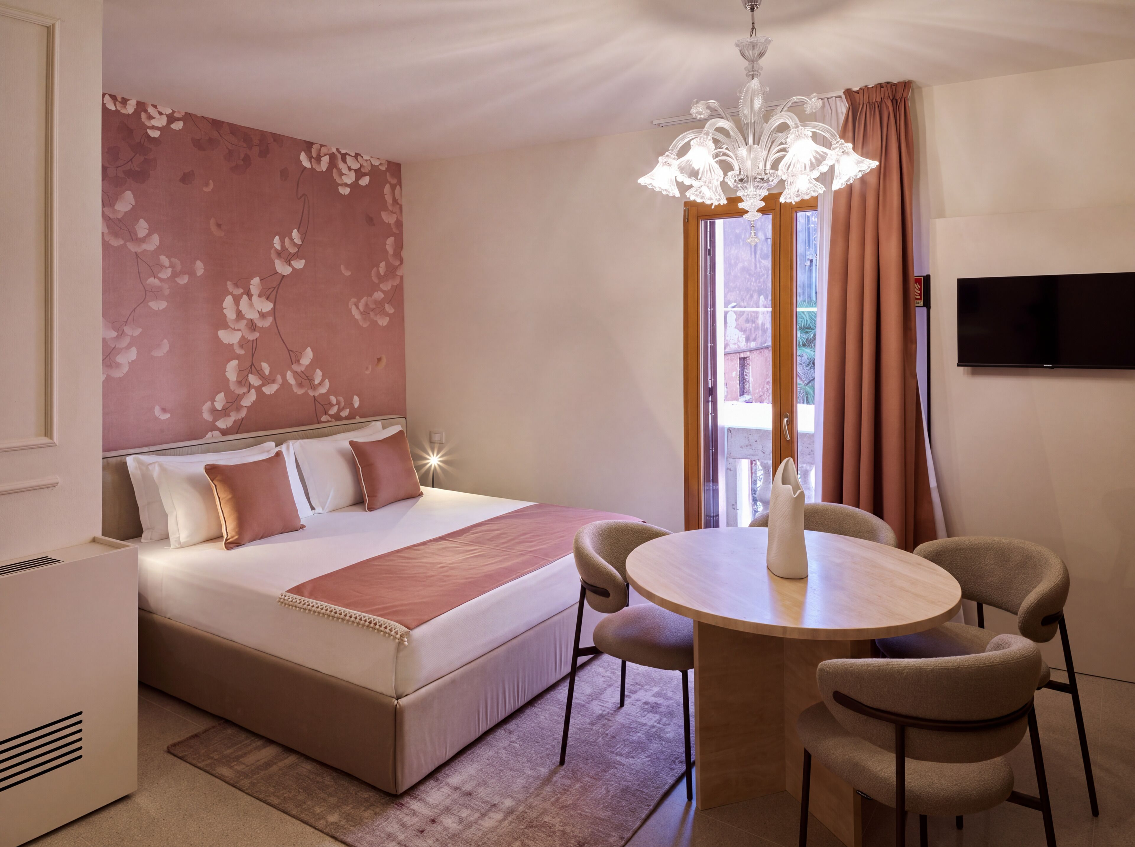 A hotel room with a bed, a round table with chairs, a television, and a chandelier.