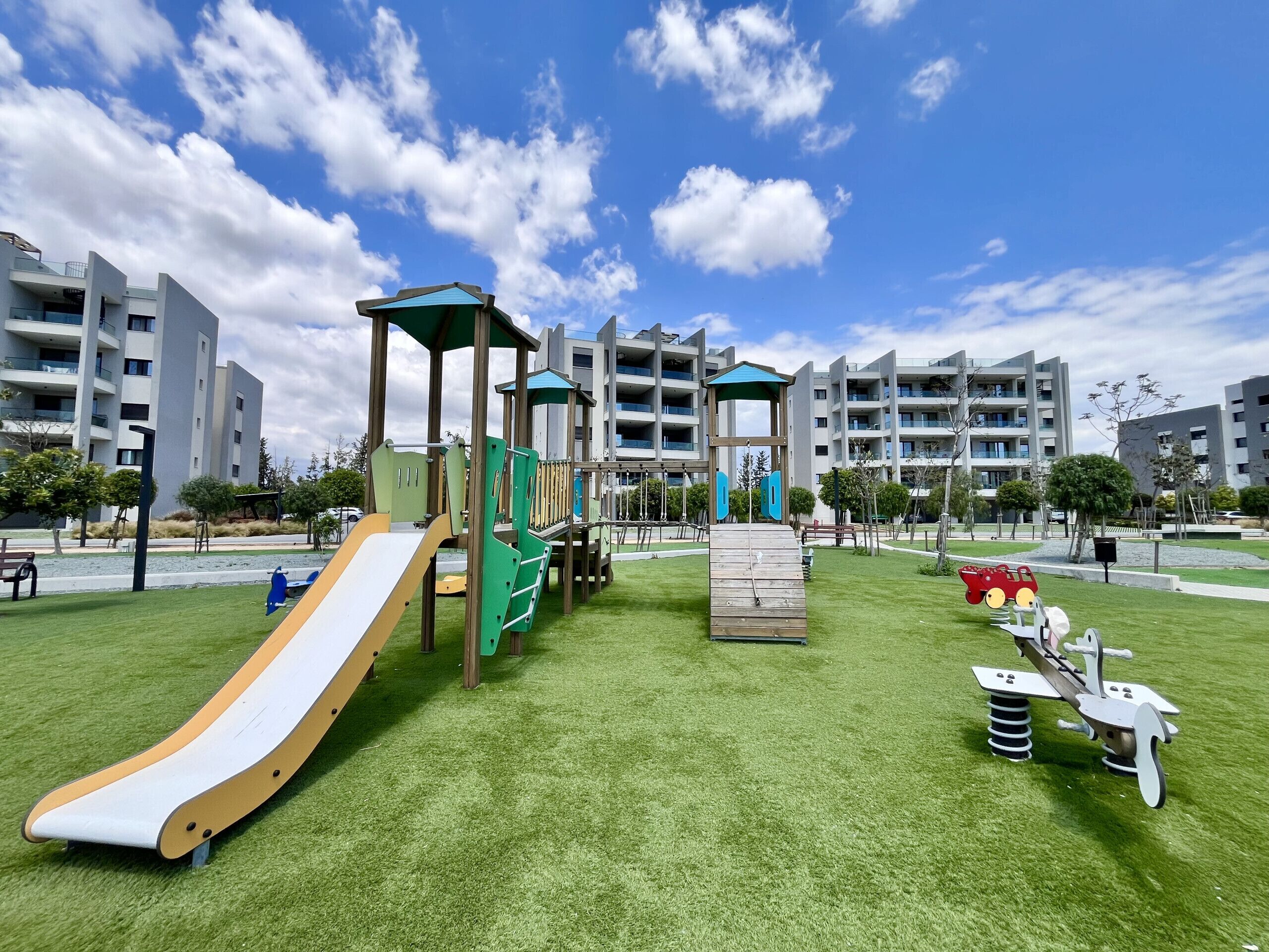 A playground with slides and climbing structures, surrounded by grass and trees, with modern buildings in the background.