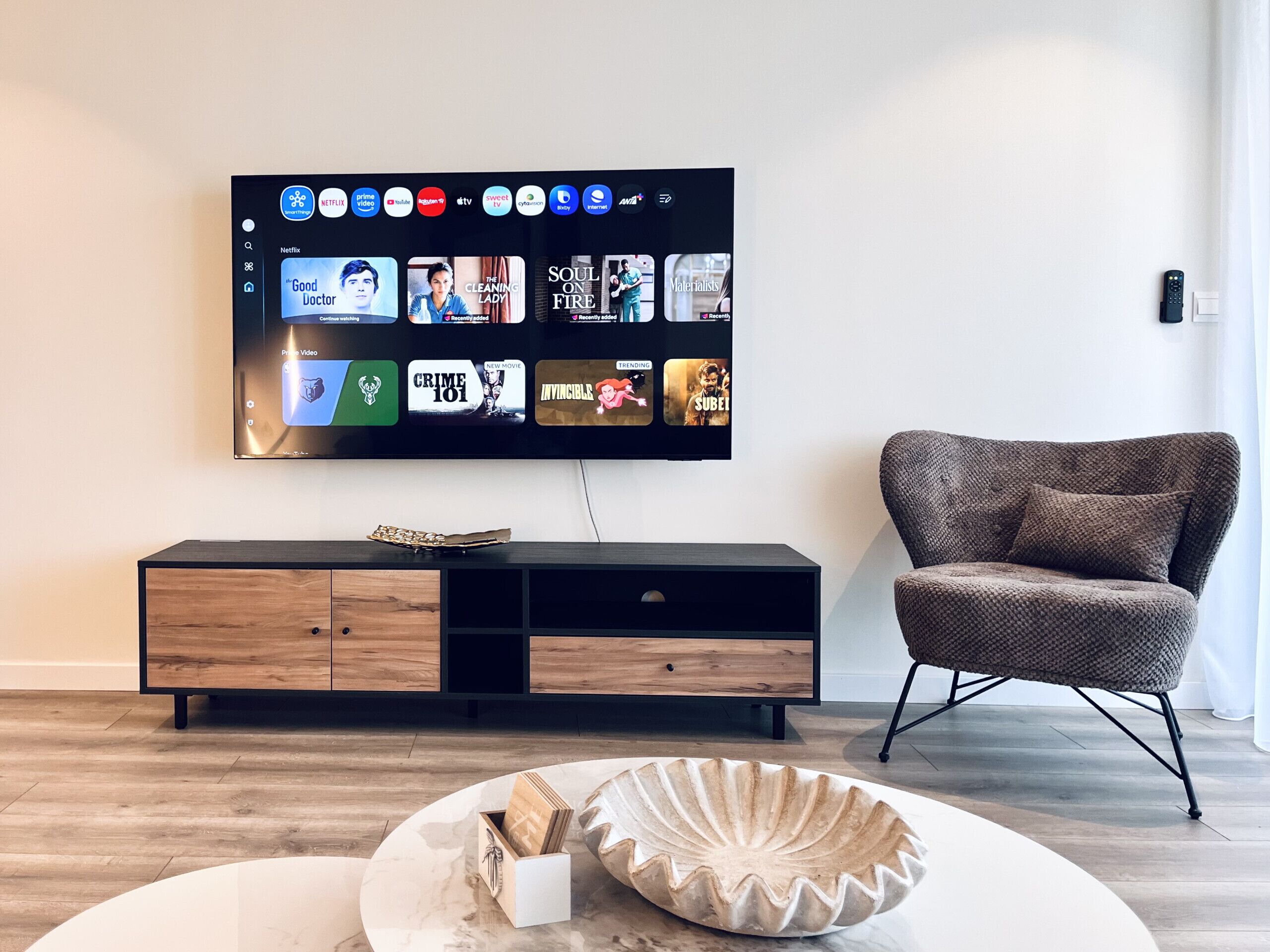 Ein modernes Wohnzimmer mit einem Flachbildfernseher, der verschiedene Streaming-Dienste anzeigt, einer Holzunterhaltungsecke, einem gemusterten Sessel und einer dekorativen Schale auf dem Boden.