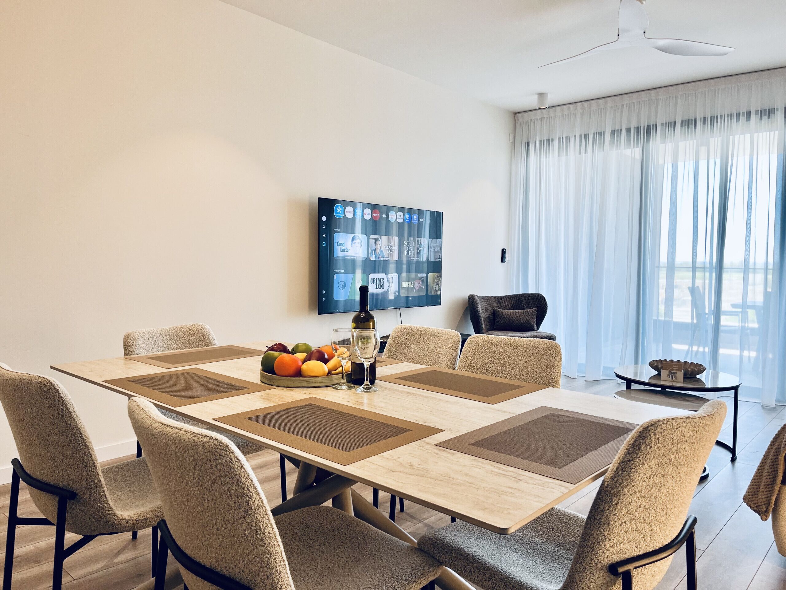 Een moderne eetruimte met een grote tafel, stoelen, een schaal fruit, een fles en een televisie aan de muur.