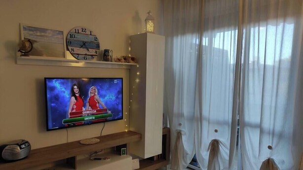 A living room with a flat-screen TV, a clock, and a window with sheer curtains. - Otta's House (Platamona)
