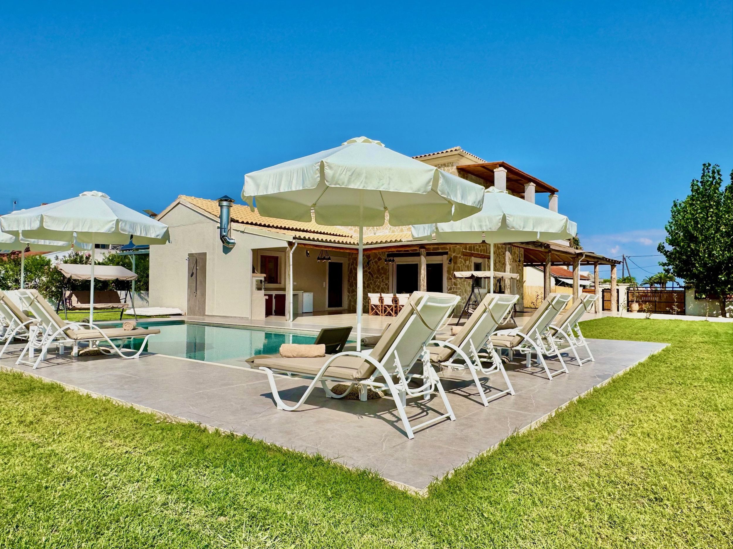 Een zwembadgedeelte met ligbedden en parasols, een overdekt terras en een stenen gebouw met pannendak.