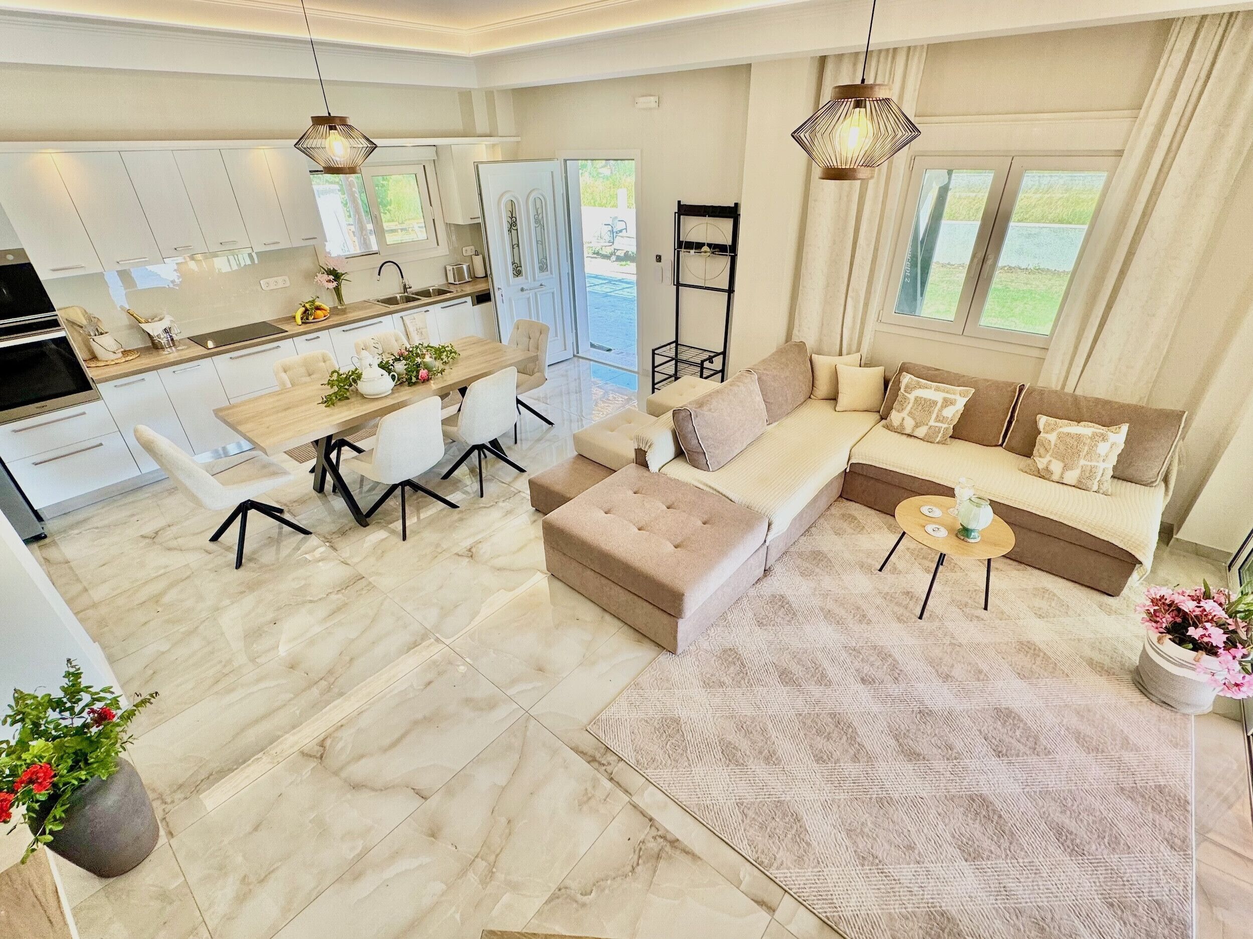 A modern living room with a sofa, ottoman, and coffee table, a dining area with a table and chairs, and a kitchen with cabinets and a window.