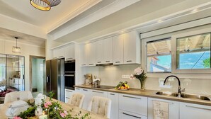 A modern kitchen with white cabinetry, a wooden dining table, and a view of the outside through a window. - Villa La Bella Sofia with private pool (Roda)