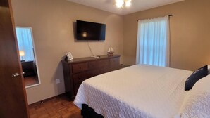 Quarto com cama, cômoda, televisão fixada na parede e janela com persianas.