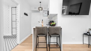 Una cocina moderna con televisor, dos taburetes de bar y una mesa pequeña con un frutero.