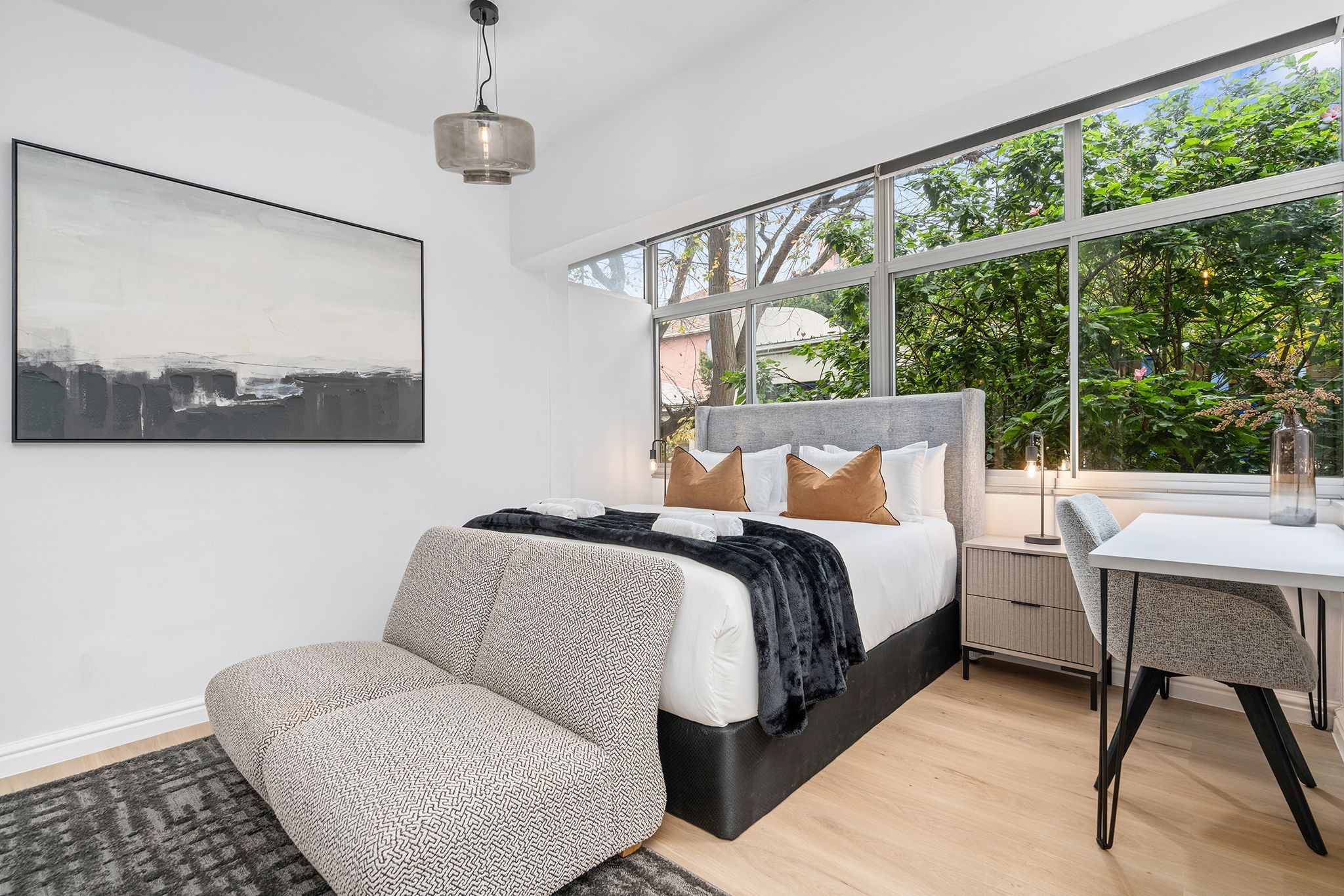 A modern bedroom with a large bed, a desk, and a view of greenery outside.