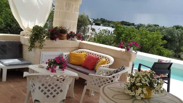 An outdoor patio with a white sofa, white chairs, a round table, and a decorative stone planter. - Villa Marica (Ostuni)