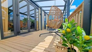 Een overdekt buitengebied met een houten terras, een hangende rieten stoel en een plant met gele vruchten.