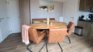 Een eetgedeelte met een houten tafel, gestoffeerde stoelen, een schilderij aan de muur en een keuken op de achtergrond.