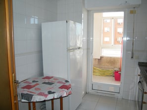 Eine kleine Küche mit weißen Fliesen, einem Kühlschrank, einer Spüle und einer Tür, die auf einen Balkon führt.