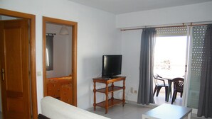 Una sala de estar con un sofá, un televisor sobre un mueble de madera, una mesa de comedor con sillas y un balcón con cortinas.