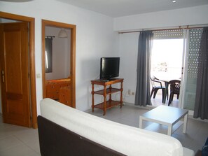 Ein Wohnzimmer mit einer Couch, einem Fernseher auf einem Holzboden, einem Esstisch mit Stühlen und einem Balkon mit Vorhängen.