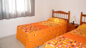 Una habitación con dos camas, una mesita de noche con lámpara, una ventana con cortinas y suelo de baldosas.