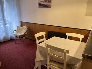 A dining area with a table and chairs, a chair with a cushion, a radiator, and a window with a curtain. - Charming studio, terrace mountain view - 4 guests (Cauterets)