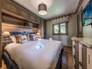 A bedroom with a wooden headboard, a bed with white linens, a bedside lamp, a window with curtains, and a TV on the wall. - Apartment chalet, relaxation - 10 guests (Courchevel)