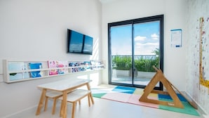 A bright room with a white table, shelves with children's items, a TV, and a large window with a view of greenery. - Corporate Stays Spirit Villa Morra (Asunción)