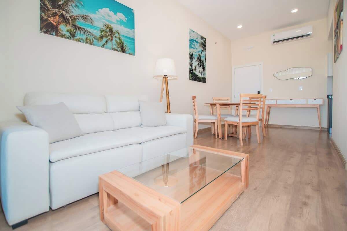 A modern living room with a white sofa, a glass coffee table, wooden chairs, and a dining table.