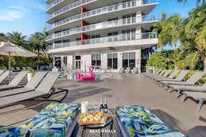 A poolside area with lounge chairs, a table with food and drinks, and a building with balconies. - Modern Studio 1BR - Bay View & Beach Access (Hallandale Beach)