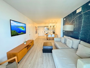A modern living room with a sofa, a TV, a wooden console, and a dining area with chairs. - Modern Studio 1BR - Bay View & Beach Access (Hallandale Beach)