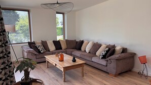 Ein modernes Wohnzimmer mit einem Ecksofa, einem Couchtisch aus Holz, einer Hängeleuchte und einem Topf mit Pflanze.