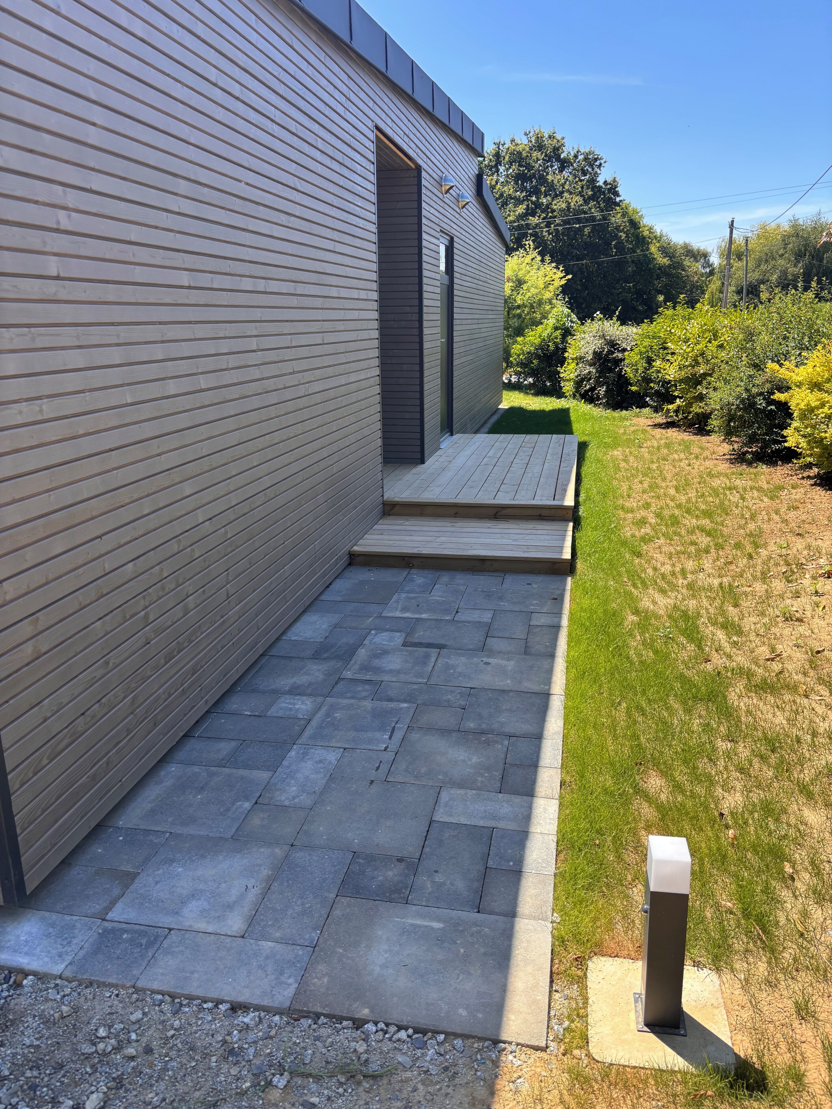 Un bâtiment moderne à l’extérieur en bois, doté d’un chemin pavé et d’une zone herbeuse agrémentée d’arbustes.