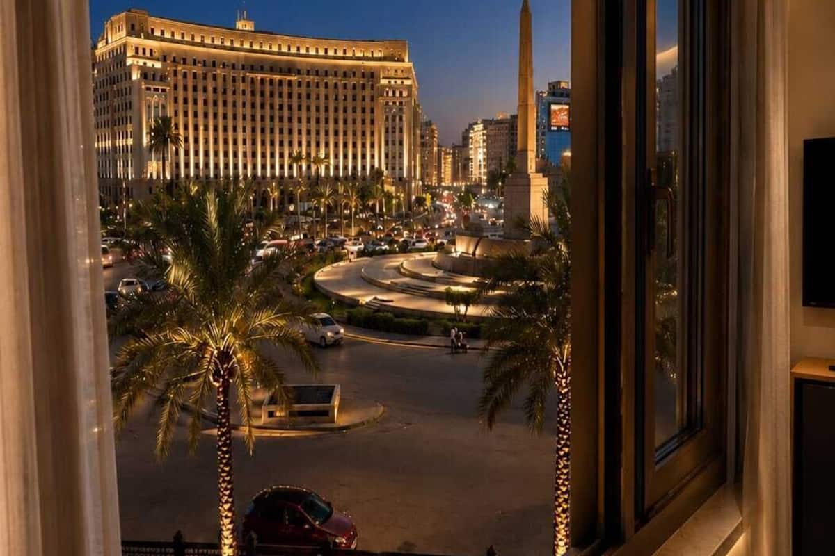 Vista desde la ventana de un hotel con vistas a un paisaje urbano que incluye un edificio grande, palmeras y un monumento.