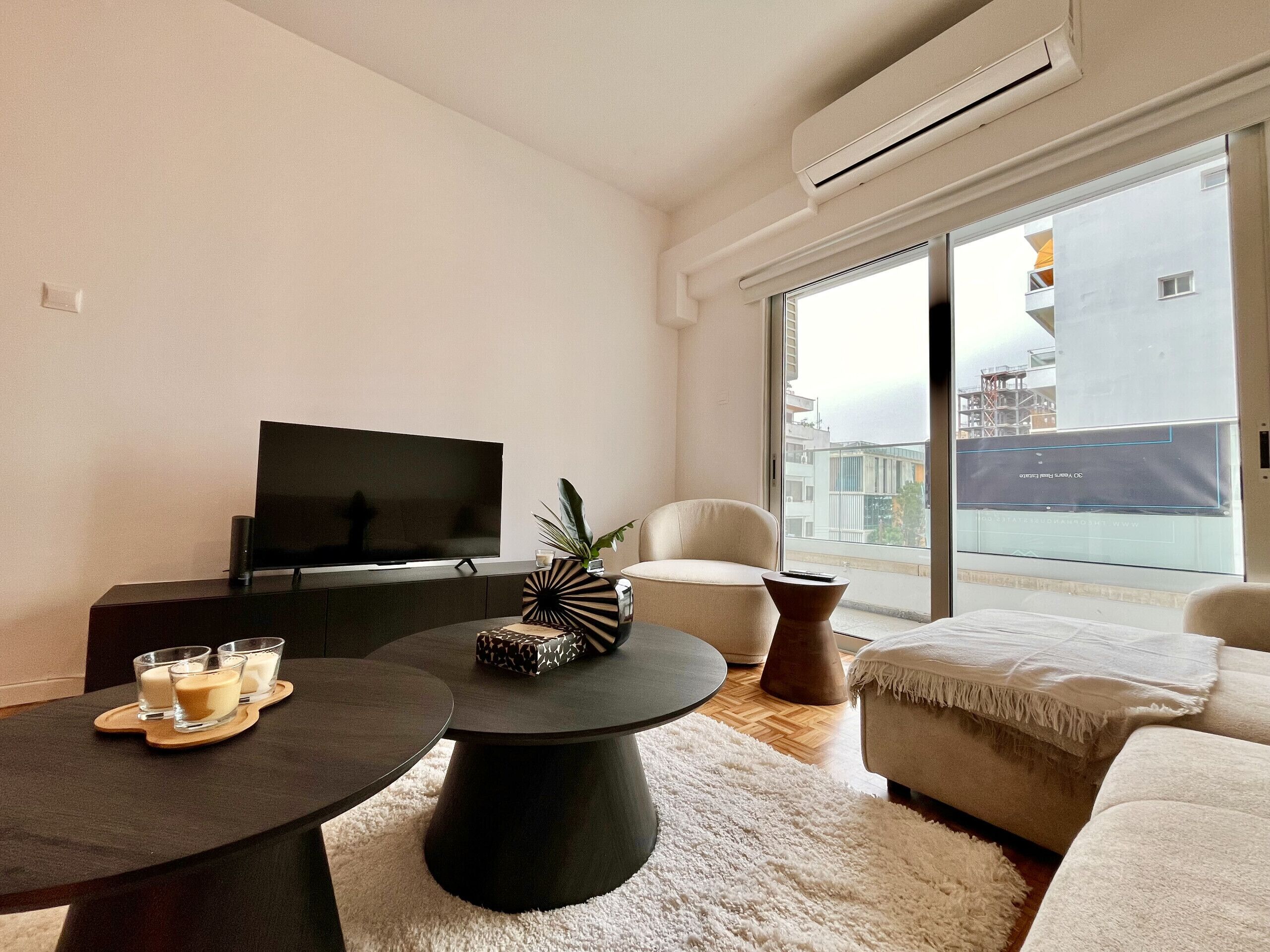 Ein modernes Wohnzimmer mit Flachbildfernseher, rundem Couchtisch, Sofa und großem Fenster mit Blick auf Gebäude.